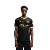 A man modeling a black Puma Manchester City soccer jersey with gold accents, featuring the Etihad Airways sponsor logo on the front, set against a plain white background.