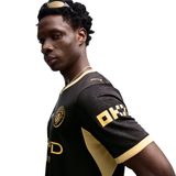 A man wearing a black and gold soccer jersey, photographed from the side against a plain white background.
