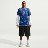Person wearing a blue patterned soccer jersey with a white long-sleeve shirt underneath, black shorts, and white sneakers on a white background.