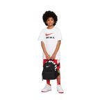 A child model wearing a white Nike t-shirt with a red swoosh and 'JUST DO IT' text, red and black patterned shorts, red socks, and white sneakers, holding a black Nike backpack against a plain white background.