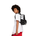 A child model wearing a white t-shirt and red shorts, shown from the side/back profile while wearing a black backpack with large white 'NIKE' lettering on the side, against a solid white background.