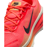 A close-up of a Nike running shoe in bright coral pink and orange, featuring a black Swoosh logo, orange laces, and a white midsole.