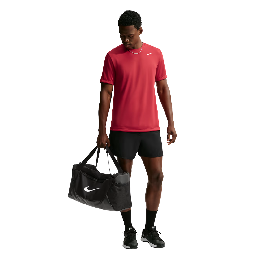 A man wearing a red Nike short-sleeve athletic shirt, black shorts, and black sneakers is shown against a white background, holding a black Nike duffel bag.