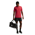 A man wearing a red Nike short-sleeve athletic shirt, black shorts, and black sneakers is shown against a white background, holding a black Nike duffel bag.