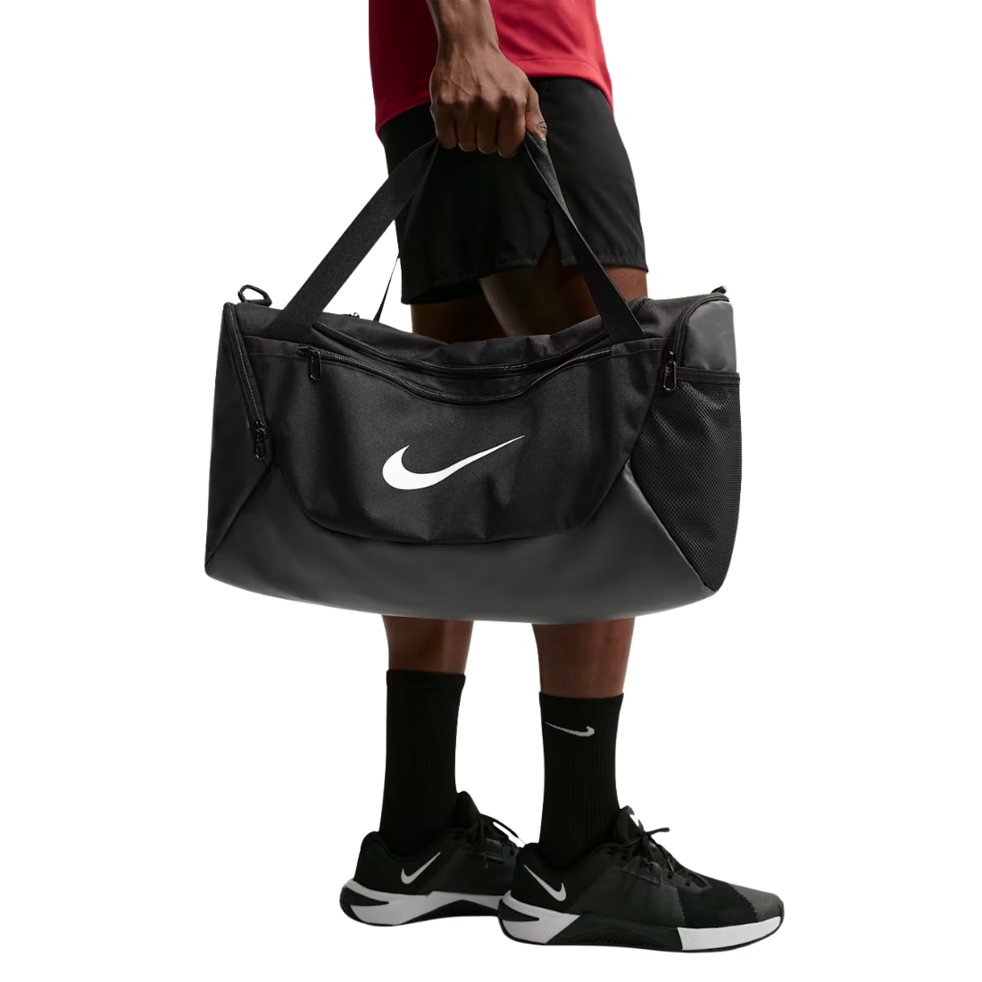 A person holding a large black Nike duffel bag with a white swoosh logo, wearing black athletic shorts, socks, and sneakers against a plain white background.