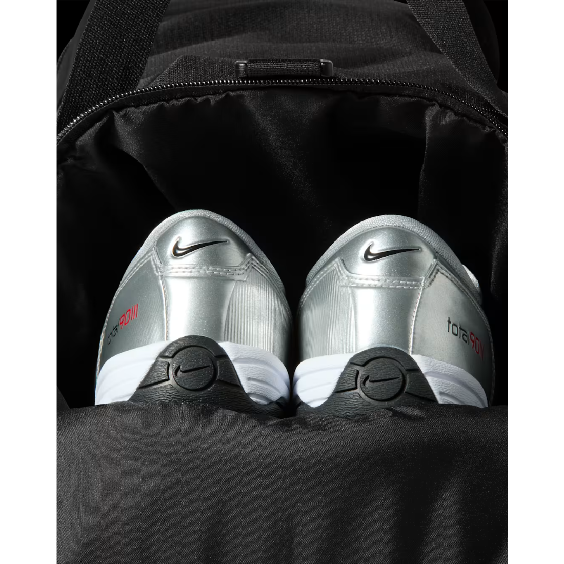 A pair of silver Nike F50 tennis shoes with black and white soles, nestled inside a black backpack.