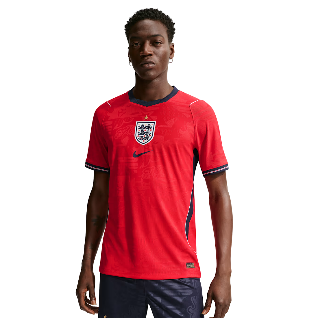 A male model wearing a red Nike England soccer jersey with navy blue trim and the England crest on the chest, paired with navy shorts, against a plain white background.