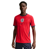 A male model wearing a red Nike England soccer jersey with navy blue trim and the England crest on the chest, paired with navy shorts, against a plain white background.