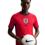 A male model wearing a red England national football team jersey by Nike, holding a white soccer ball against a solid white background.