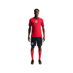 A male model wearing a red England national football team home kit by Nike, featuring a red jersey with the England crest and Nike swoosh, navy shorts, and red socks, set against a plain white background.