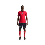 A male model wearing a red England national football team home kit by Nike, featuring a red jersey with the England crest and Nike swoosh, navy shorts, and red socks, set against a plain white background.