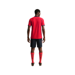 A man viewed from the back wearing a red short-sleeve soccer jersey, navy shorts, and red socks against a plain white background.