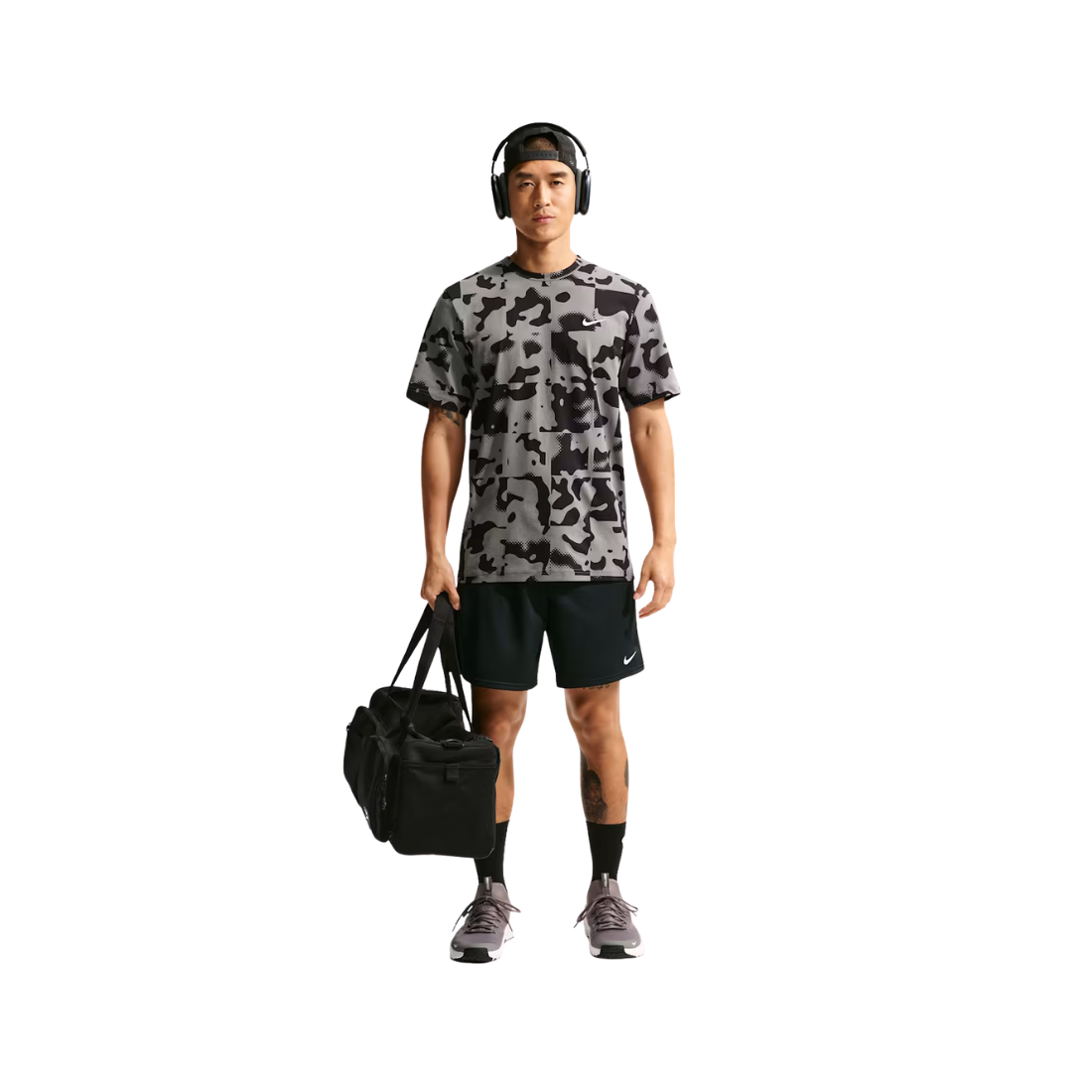 A man wearing a gray and black camouflage t-shirt, black athletic shorts, and headphones stands against a white background, holding a black duffel bag.