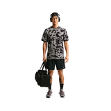 A man wearing a gray and black camouflage t-shirt, black athletic shorts, and headphones stands against a white background, holding a black duffel bag.