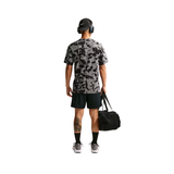 A man shown from the back wearing a gray and black camouflage short-sleeve athletic t-shirt, black shorts, black socks, and gray sneakers, while holding a black duffel bag against a plain white background.
