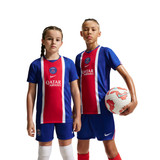 Two children modeling Paris Saint-Germain soccer jerseys and shorts against a white studio background. The girl wears a blue and red jersey with white vertical stripes, and the boy wears a matching blue and red jersey with white vertical stripes.