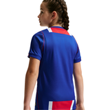 A child model shown from the back wearing a royal blue short-sleeve athletic jersey with red and white geometric accents on the shoulders and lower back, against a plain white studio background.