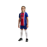 A young girl modeling a Paris Saint-Germain soccer kit, featuring a blue and red jersey with white stripes, matching blue shorts, white knee-high socks, and black Nike cleats, set against a plain white background.