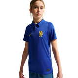 A young girl modeling a royal blue short-sleeve polo shirt with a yellow Nike swoosh and a yellow Chelsea FC lion crest on the chest, set against a plain white background.