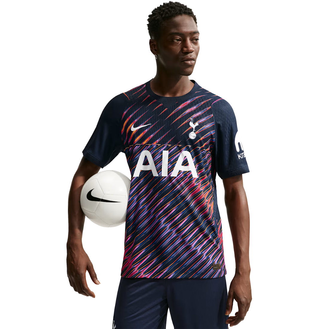 A male athlete wearing a navy blue and pink patterned Tottenham Hotspur soccer jersey with the white AIA sponsor logo, holding a white soccer ball against a solid white background.