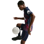 A soccer player in a navy blue and red striped jersey and shorts, balancing a white soccer ball on his foot against a solid white background. The jersey features a visible 'Kraken' sponsor logo on the sleeve.