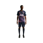 A male model wearing a Tottenham Hotspur soccer kit, featuring a navy blue jersey with a diagonal pink and purple pattern, navy shorts, and navy socks, on a solid white background.