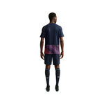 A male model shown from the back wearing a navy blue soccer kit, featuring a short-sleeve jersey with a pink and purple patterned band at the waist, matching shorts, and navy socks.