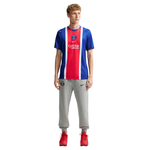 A mannequin wearing a Paris Saint-Germain Nike home soccer jersey in blue, red, and white, paired with gray sweatpants and red sneakers, on a solid white background.