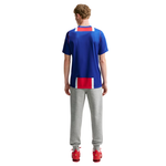 A man viewed from behind wearing a royal blue athletic t-shirt with red and white block accents, paired with gray sweatpants and red sneakers, against a plain white background.