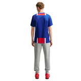 A man viewed from behind wearing a royal blue athletic t-shirt with red and white block accents, paired with gray sweatpants and red sneakers, against a plain white background.