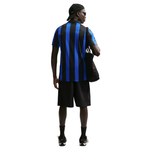 A man shown from the back wearing a black and blue vertically striped short-sleeve soccer jersey, black athletic shorts, a black cap, sunglasses, and carrying a black shoulder bag against a plain white background.