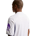A man shown from the back wearing a white short-sleeve athletic jersey with purple side panels and a purple logo on the sleeve, photographed against a plain white background.