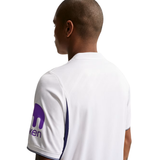 A man shown from the back wearing a white short-sleeve athletic jersey with purple side panels and a purple logo on the sleeve, photographed against a plain white background.