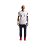 A male model stands against a solid white background wearing a white Tottenham Hotspur Nike soccer jersey with red AIA sponsor text, navy blue pants, and red soccer cleats.