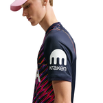 A side-profile view of a male model wearing a navy blue athletic jersey with a colorful diagonal stripe pattern on the lower front and a white Kraken logo on the sleeve, paired with a pink cap against a white background.