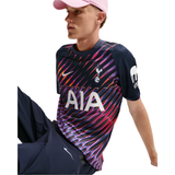 A model wearing a navy blue Tottenham Hotspur soccer jersey with a vibrant pink, purple, and orange wavy stripe pattern, paired with navy shorts and a pink cap against a white background.