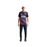 A man modeling a navy blue Tottenham Hotspur athletic t-shirt with a pink and purple geometric pattern, paired with navy pants, black sneakers, and a pink cap, against a solid white background.