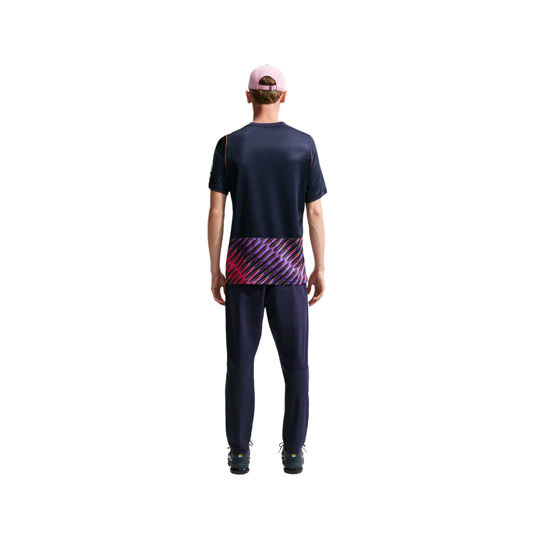 A man viewed from behind wearing a dark navy athletic t-shirt with a pink and purple patterned waistband, matching navy athletic pants, and a pink cap, set against a plain white background.