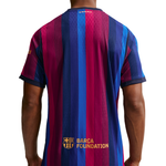 Back view of a short-sleeve soccer jersey worn by a model, featuring vertical stripes in magenta, royal blue, and navy, with the FC Barcelona crest and 'BARCA FOUNDATION' text near the lower back.