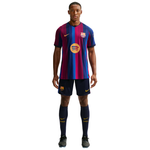 A man modeling a Barcelona soccer kit consisting of a short-sleeve jersey with blue and magenta vertical stripes, navy shorts, and navy socks, on a plain white background.