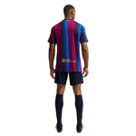 A man viewed from the back wearing a short-sleeve soccer jersey with vertical blue, magenta, and navy stripes, navy shorts, and navy knee-high socks on a white background.