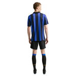 A man shown from the back wearing a short-sleeve soccer jersey with vertical blue and black stripes, black shorts, and black knee-high socks with blue bands, against a plain white background.