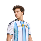 A man wearing an Argentina national football team home jersey by Adidas, featuring light blue and white vertical stripes with gold details, set against a plain white background.