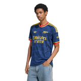 A man wearing a blue Arsenal football jersey with yellow and red accents, paired with light blue jeans, photographed against a plain white background.