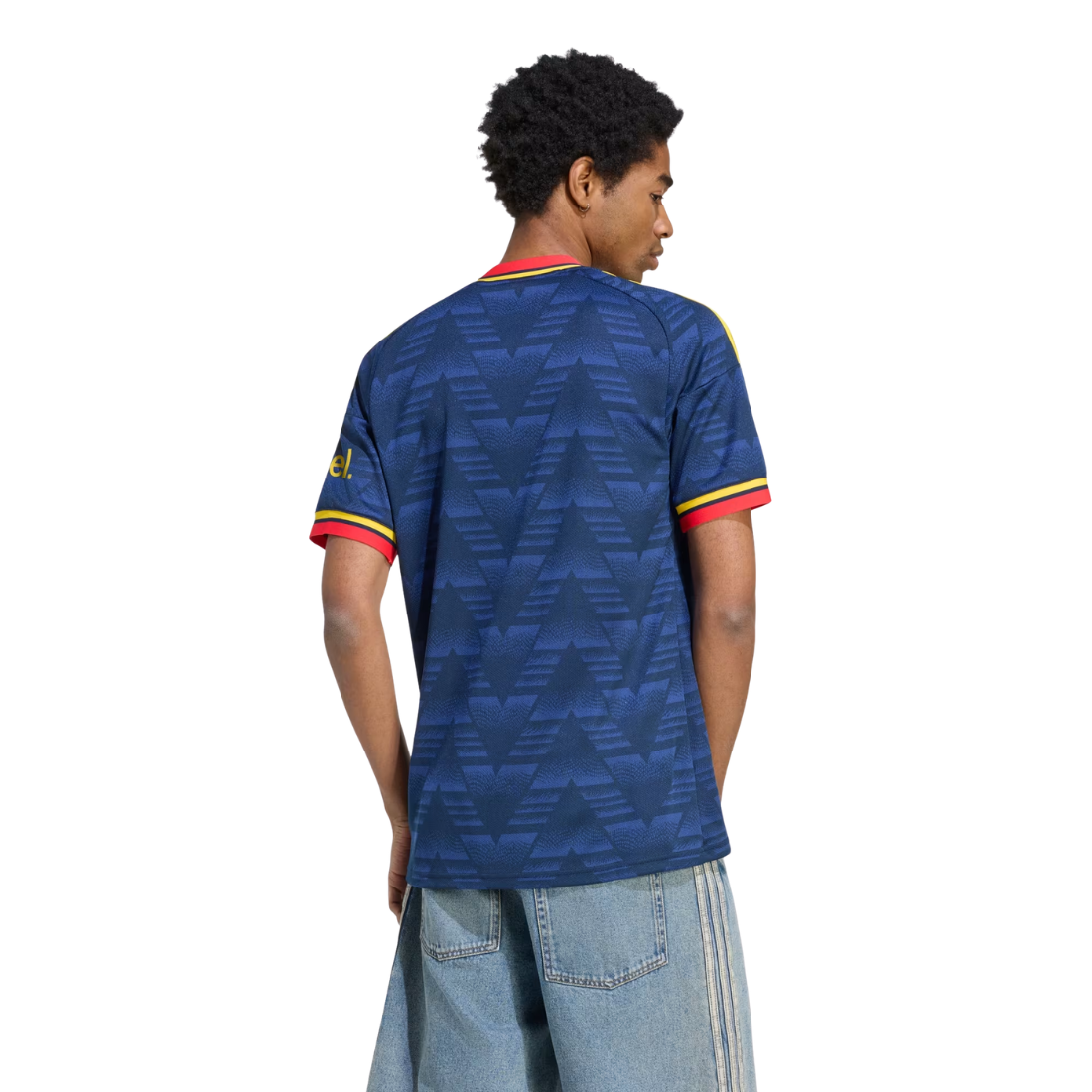 A man shown from the back wearing a blue patterned soccer jersey with red and yellow trim on the collar and sleeve cuffs, paired with light blue jeans, against a plain white background.