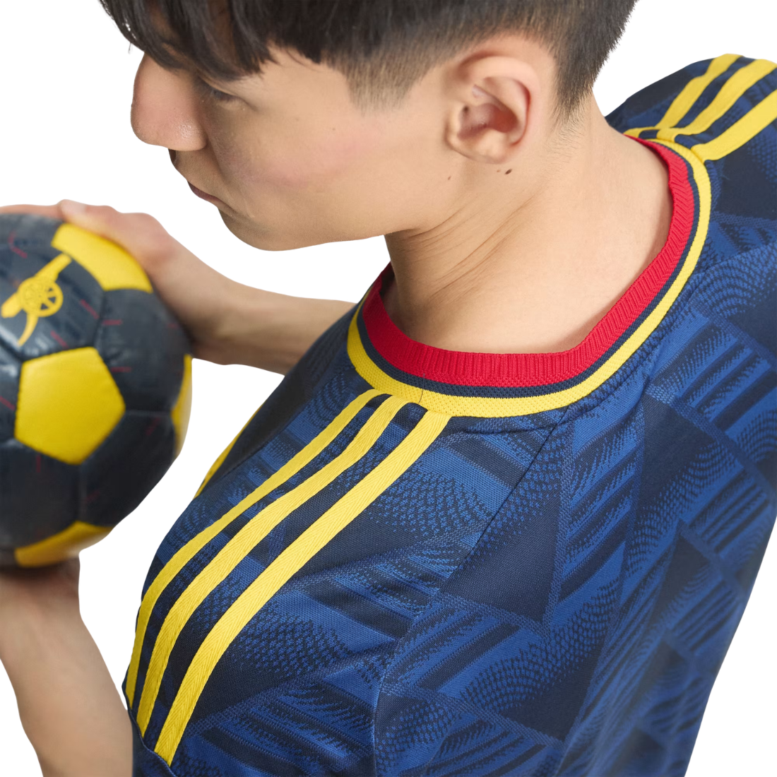 A close-up side profile of a person wearing a blue soccer jersey with yellow stripes and a red collar, holding a black and yellow soccer ball against a white background.