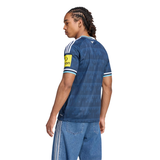 A man shown from the back wearing a blue Adidas soccer jersey with white stripes on the shoulders and sleeves, paired with blue jeans with white stripes down the sides, against a plain white background.