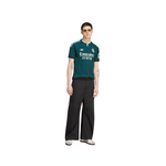 A model wearing a dark green Real Madrid soccer jersey with white accents, paired with black wide-leg trousers and white sneakers, set against a plain white background.