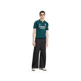 A model wearing a dark green Real Madrid soccer jersey with white accents, paired with black wide-leg trousers and white sneakers, set against a plain white background.