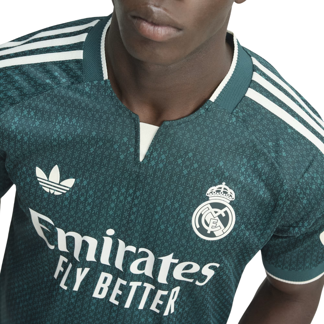 A close-up of a person wearing a Real Madrid home soccer jersey by Adidas. The jersey is dark teal green with a subtle textured pattern, featuring white stripes on the shoulders, white sponsor text 'Emirates FLY BETTER', the Adidas Trefoil logo, and the Real Madrid crest.